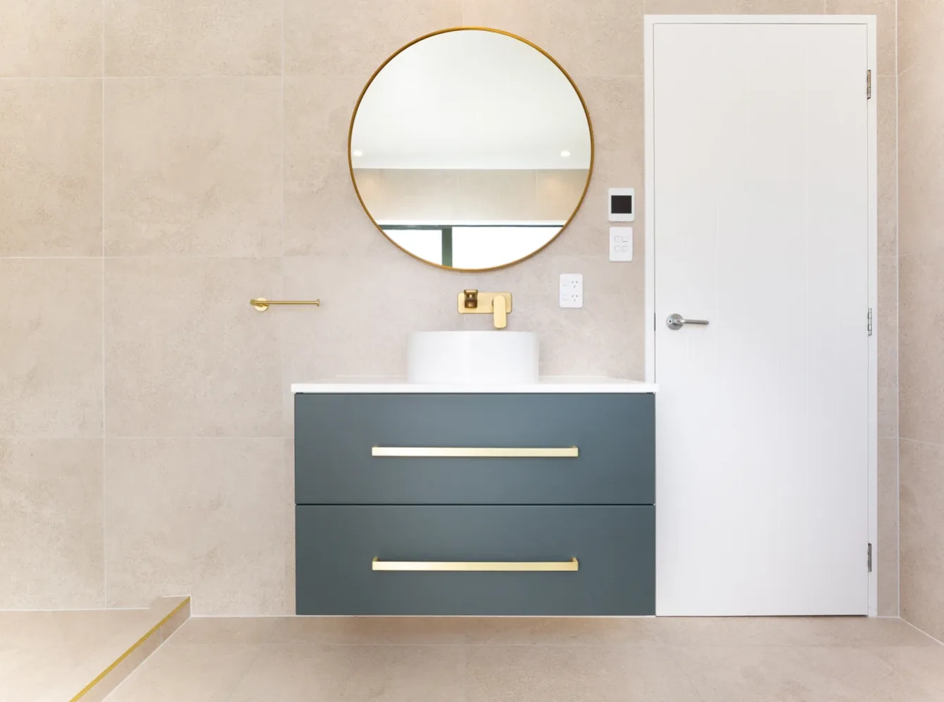 Dark teal wall-hung vanity with two drawers and long gold handles, a white round vessel basin, gold wall-mounted mixer, and a large round gold-framed mirror above.