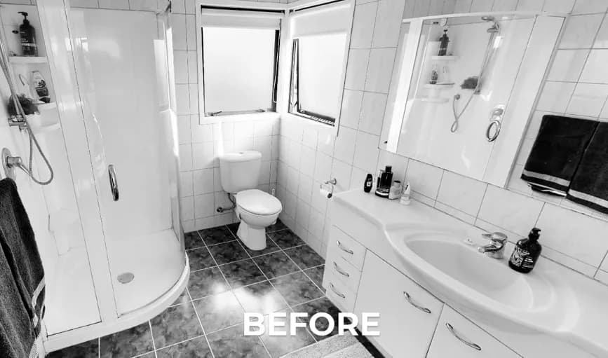 Black-and-white photograph of the bathroom before renovation, showing a corner shower enclosure, a wall-hung toilet beneath a window, and a laminate vanity with a mirrored cabinet.