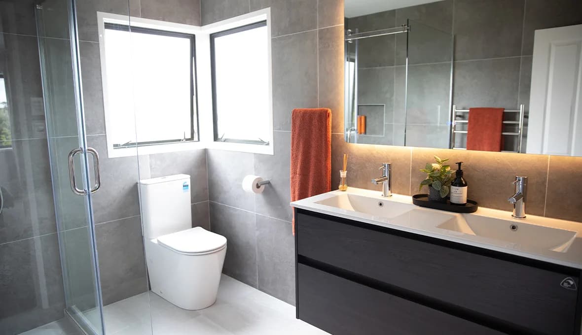 Bathroom with grey stone-look tiles, a double vanity with two basins and matte black tapware, a wall-hung toilet in a corner window nook, a terracotta towel on a heated rail, and a frameless glass shower.