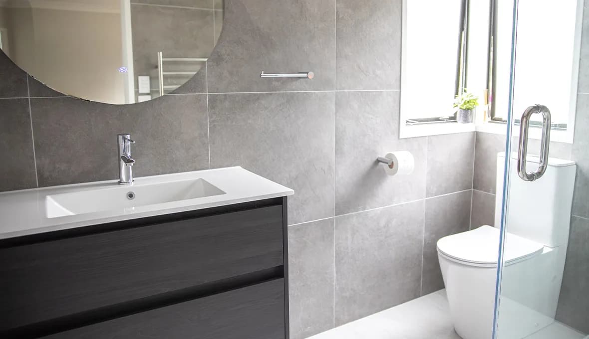 Ensuite with a single vanity, a round mirror, chrome tapware, a wall-hung toilet behind a frameless glass shower screen, and a small window with a potted plant on the sill.