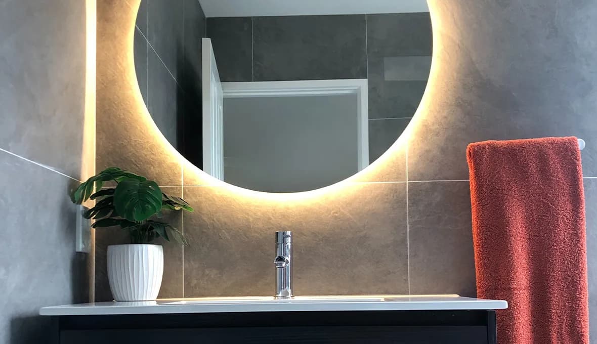 Round LED backlit mirror above a dark vanity with a potted plant, reflecting a doorway, set against charcoal stone-look tiles.