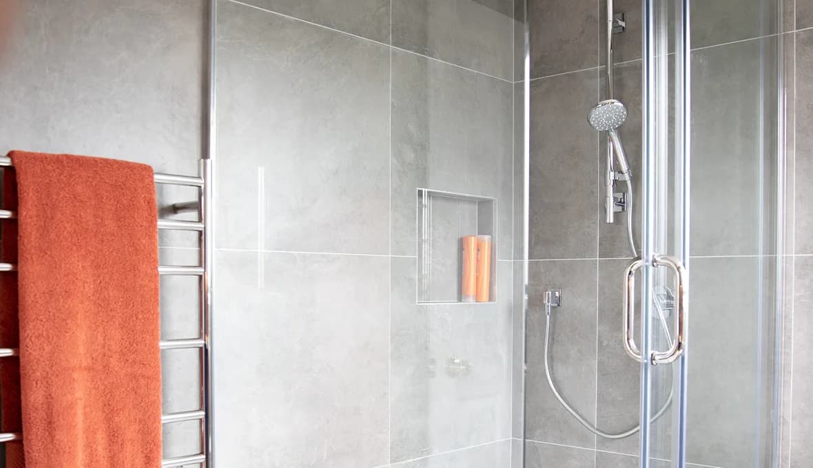 Chrome rail shower with a handheld head in a frameless glass shower, beside a recessed tiled niche holding two terracotta bottles.