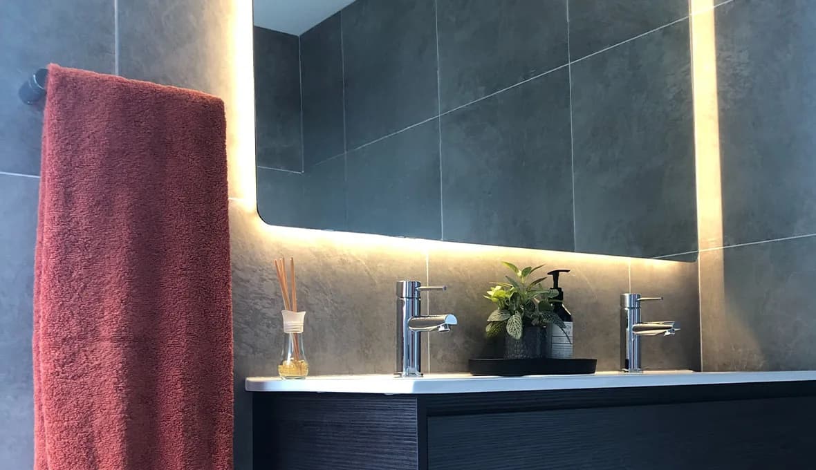 Close-up of a chrome basin tap on a dark vanity, with a terracotta towel, a reed diffuser and a potted plant beside a backlit mirror glowing warm light.