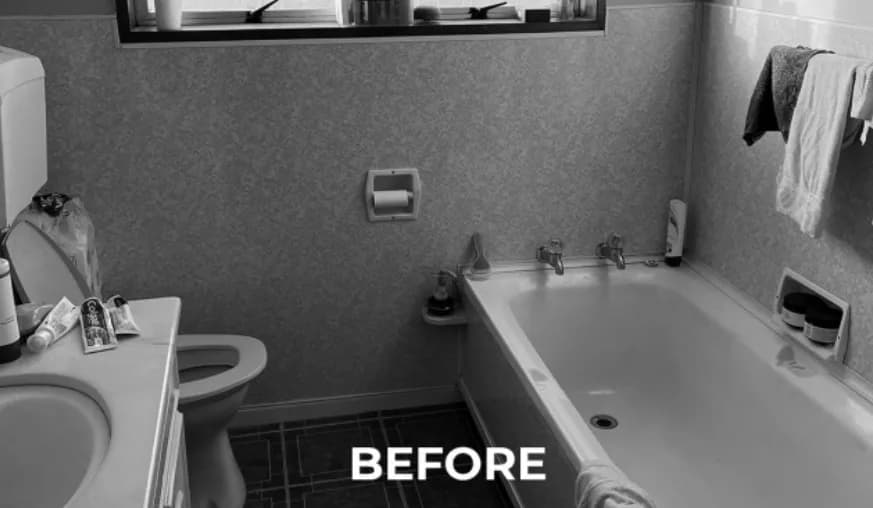 Black-and-white photograph of the bathroom before renovation, showing textured cream wall tiles, dark patterned floor tiles, a built-in bath with two chrome taps, and a white vanity unit beside the toilet.