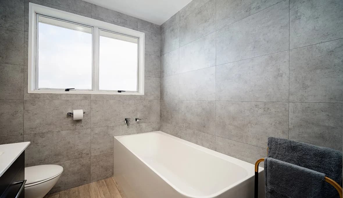 White built-in bath below a window, with a chrome wall-mounted mixer, grey concrete-look tiles, timber-look floor, and a ladder-style towel rail holding two dark grey towels.