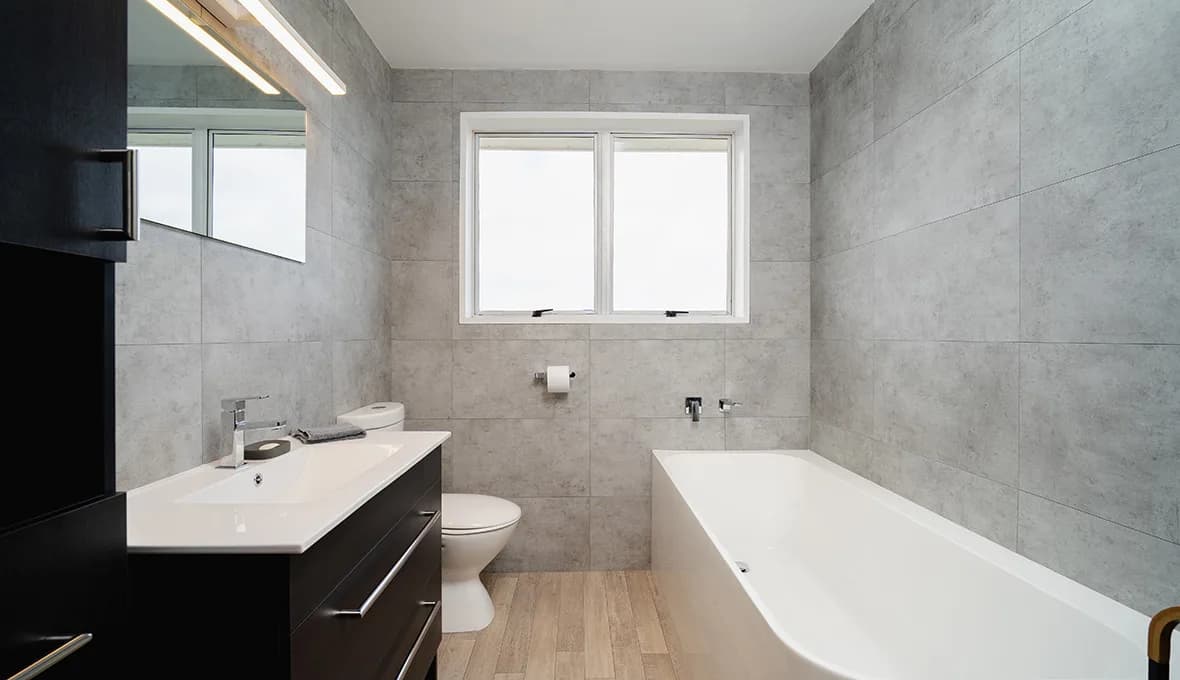 Bathroom with grey concrete-look wall tiles and timber-look floor tiles, a dark timber wall-hung vanity with a white top and chrome tap, a wall-hung toilet, and the foot of a white built-in bath in the foreground.
