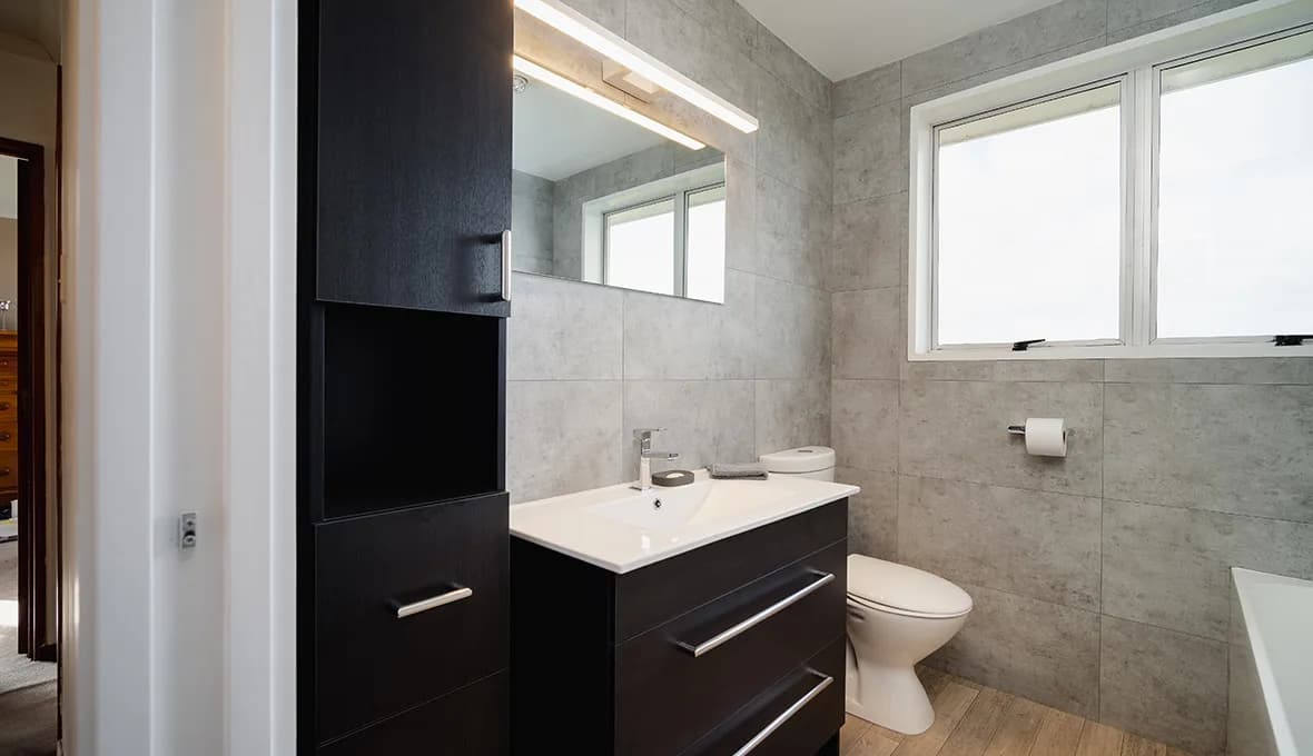 Dark timber tall storage cabinet beside a matching wall-hung vanity with a white top and chrome tap, a wall-hung toilet, and a window set into grey concrete-look wall tiles.