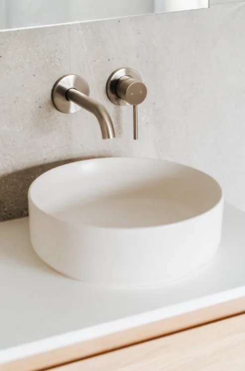 Close-up of a round white vessel basin on a white benchtop with a brushed nickel wall-mounted spout and mixer against speckled stone-look tiles.