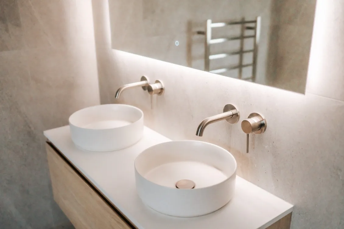 Angled close view of two round white vessel basins on a white benchtop, with brushed nickel wall-mounted spouts and mixers and an LED backlit mirror reflecting a heated towel rail.