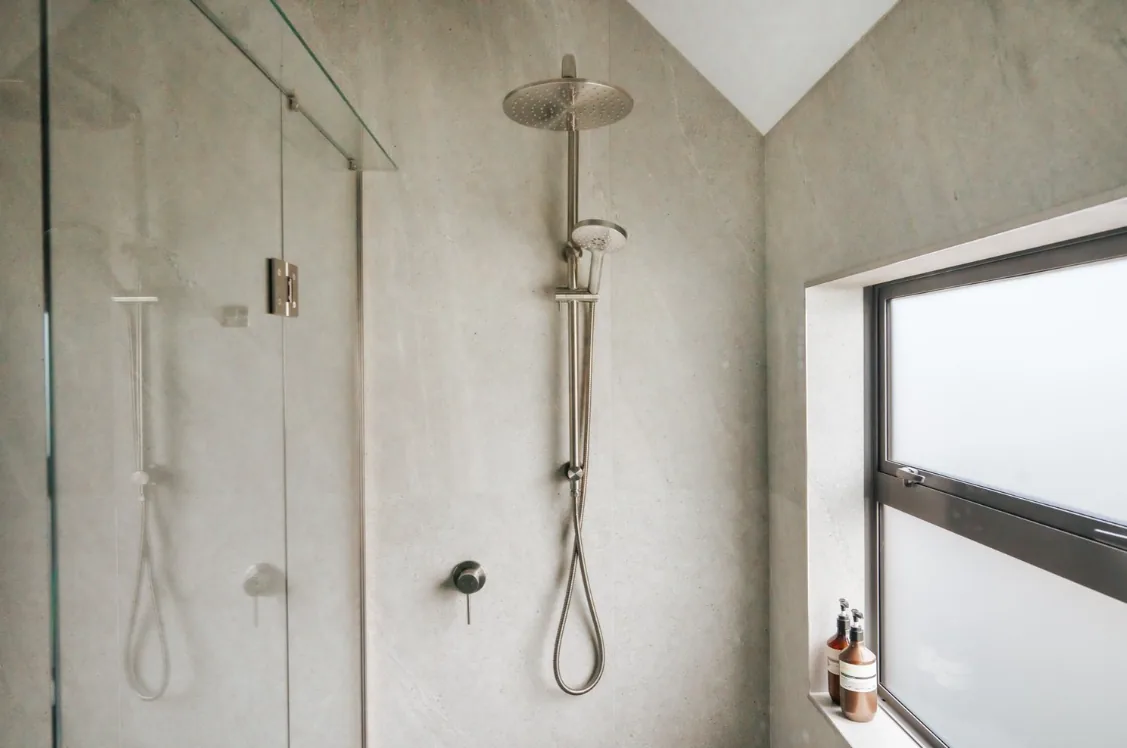 Shower area with a brushed nickel rain head on a riser rail with a handheld shower, a frameless glass screen, and an obscured-glass window with two amber bottles on the sill.