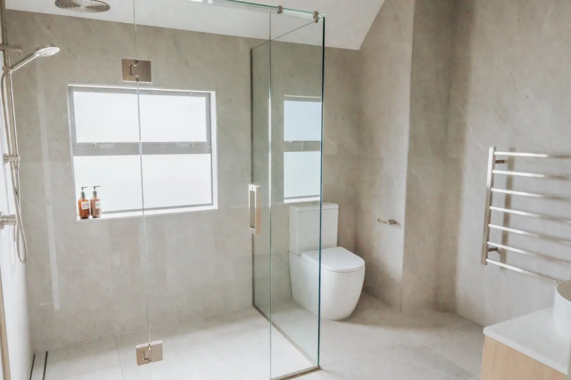 Frameless glass shower enclosure with a rain head and handheld rail, beside a close-coupled white toilet and a stainless steel ladder-style heated towel rail, all against large-format grey stone-look tiles.