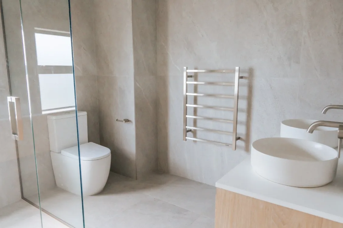 White close-coupled toilet viewed past the edge of a frameless glass shower screen, with a stainless steel ladder heated towel rail on the wall and a light oak vanity with a round basin in the foreground.