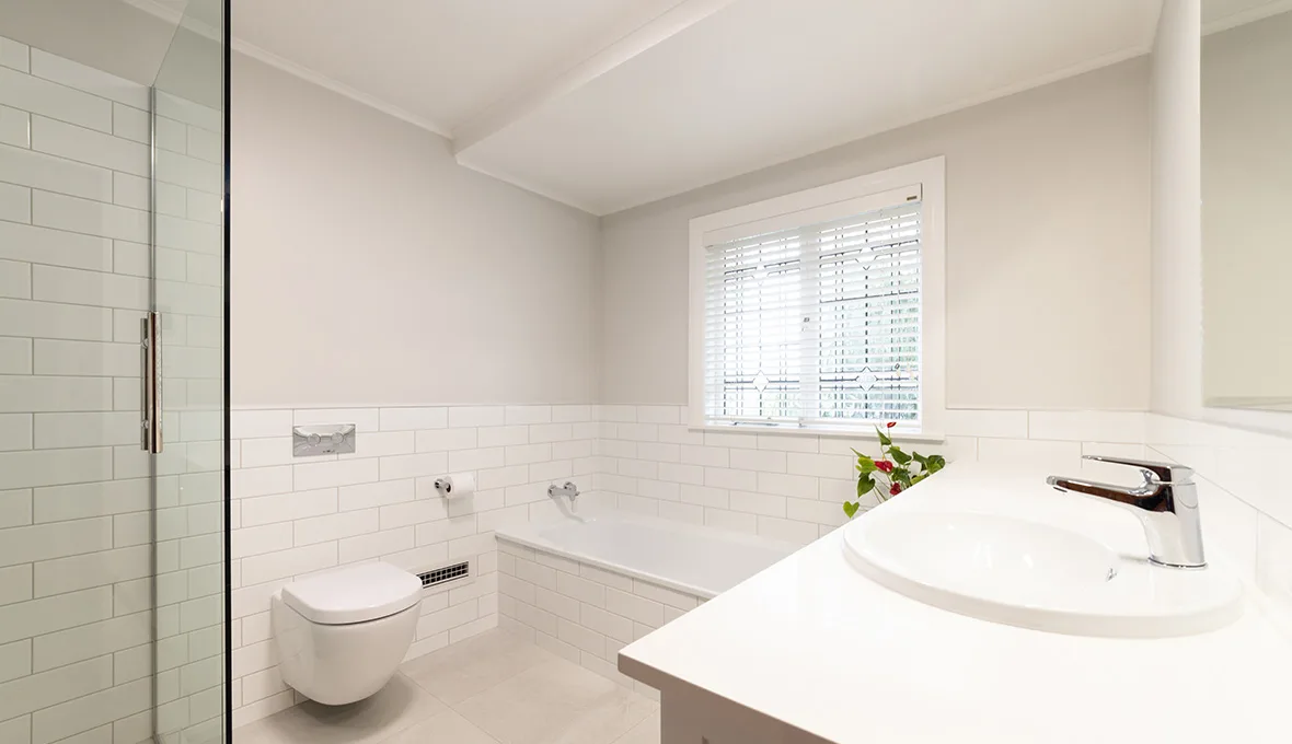 White bathroom with subway-tile walls, a frameless glass shower screen, a wall-hung toilet, a built-in bath below a window with patterned blinds, and the edge of a white vanity with a chrome tap in the foreground.