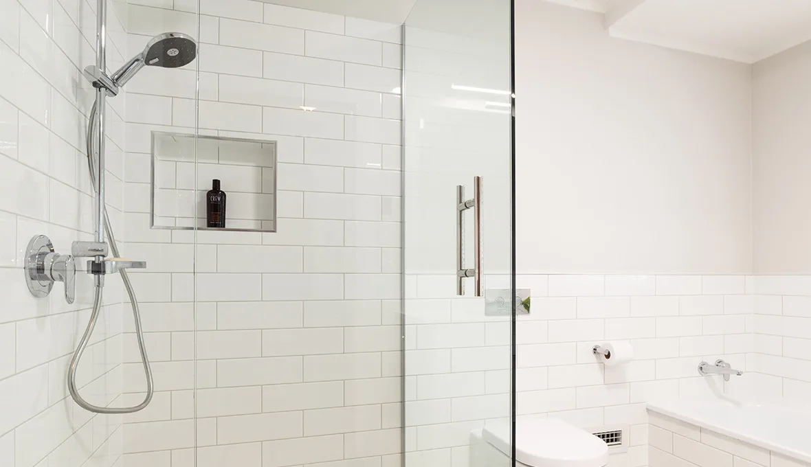 Corner of a frameless glass shower with a chrome rail shower and handheld head, a recessed tiled niche holding a dark bottle, and white subway tile walls.