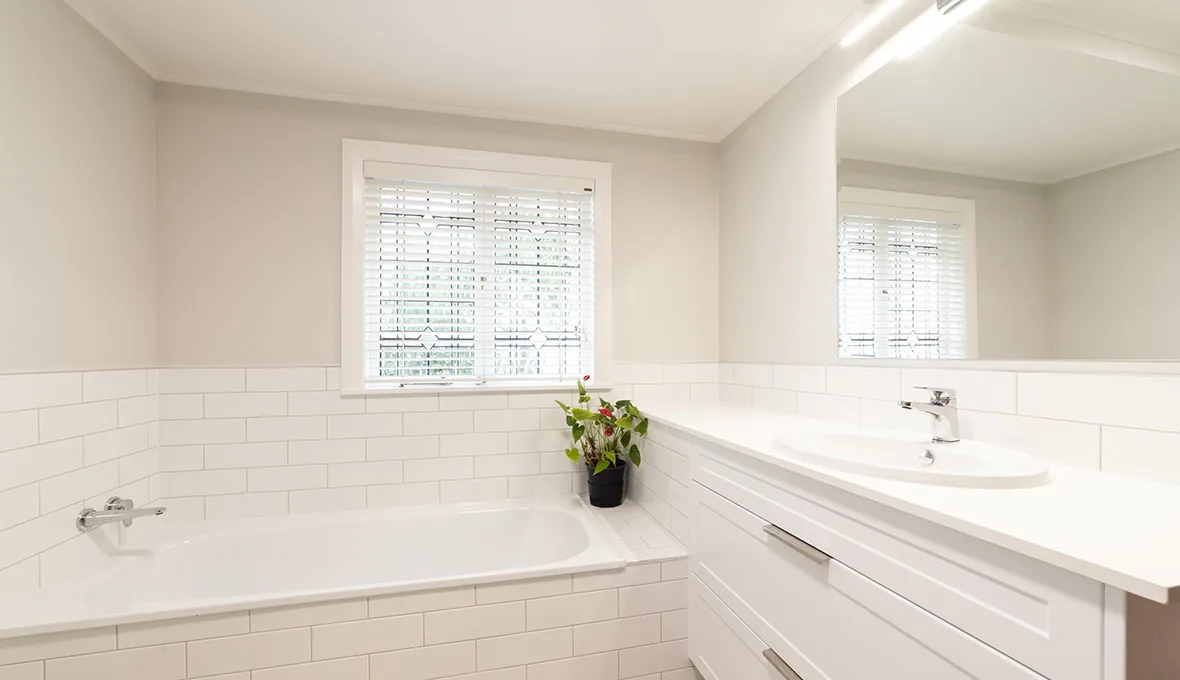 White built-in bath and a white vanity with a chrome tap, reflected in a wall mirror, with white subway tile walls and a window with patterned blinds.