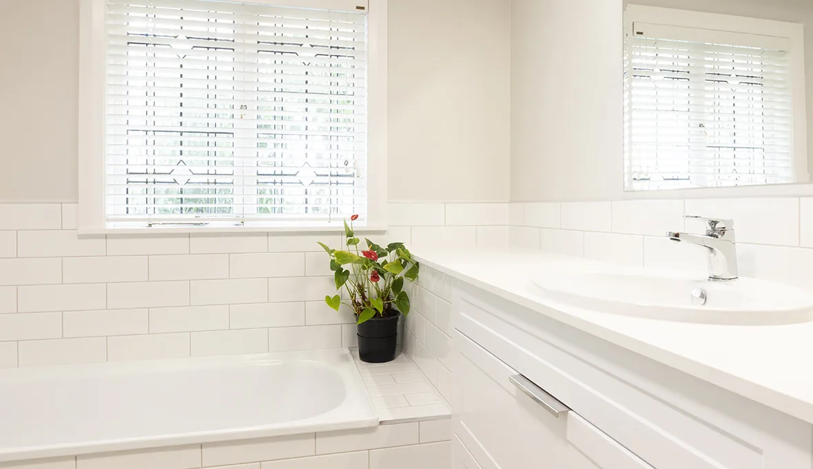 White built-in bath below a window with patterned blinds, a potted plant with red flowers on the tiled ledge, and the edge of a white vanity with a chrome tap.