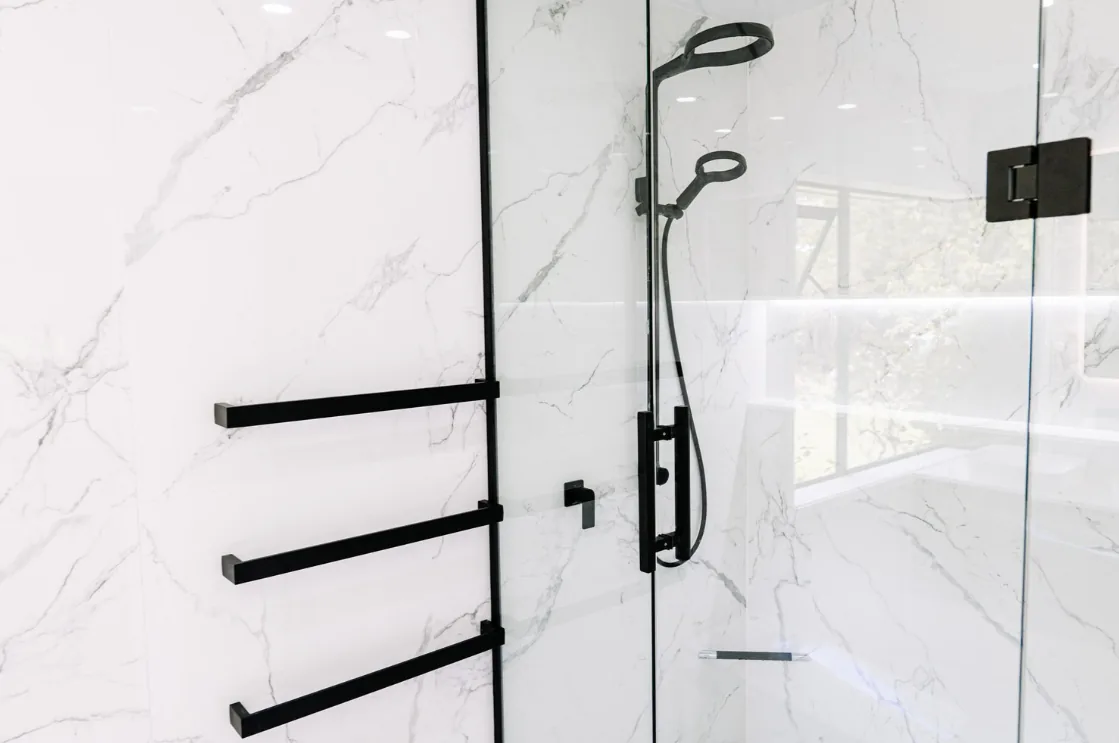 Three black towel rails mounted on white marble-look tiling beside a frameless glass shower with a black rain head, handheld shower and black wall mixer.
