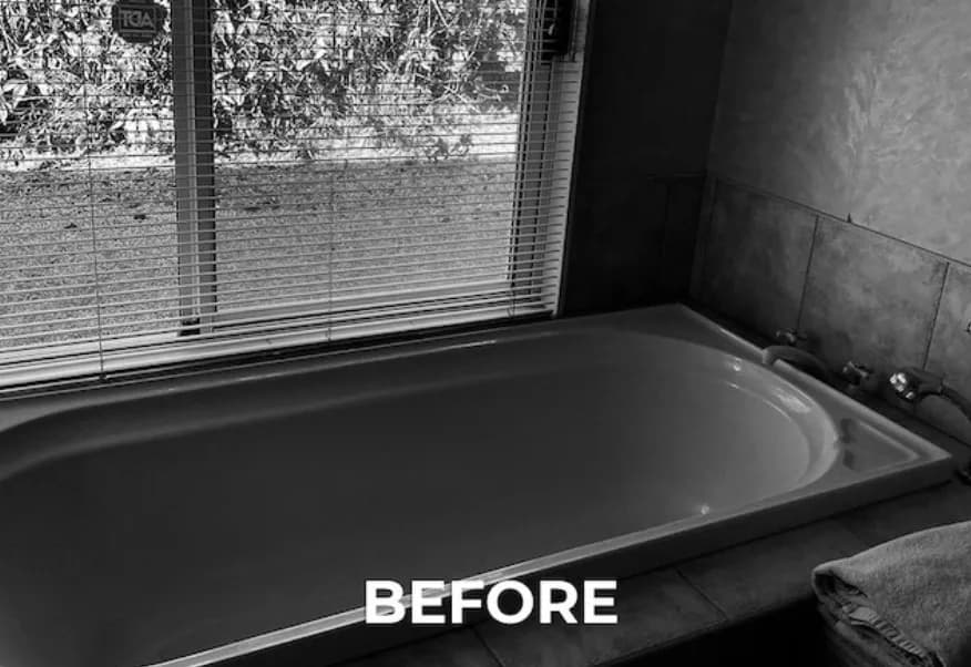 Black-and-white photograph of the bathroom before renovation, showing a dated built-in bath with two chrome taps and a tiled surround, viewed through a window with venetian blinds.