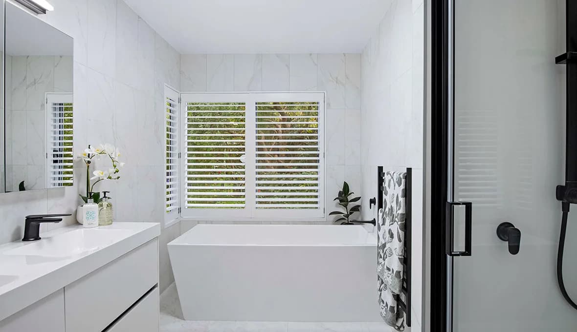 White freestanding bath beneath timber plantation shutters looking onto greenery, with full-height white marble-look wall tiles, a matte black ladder towel rail, and the edge of a frameless glass shower.