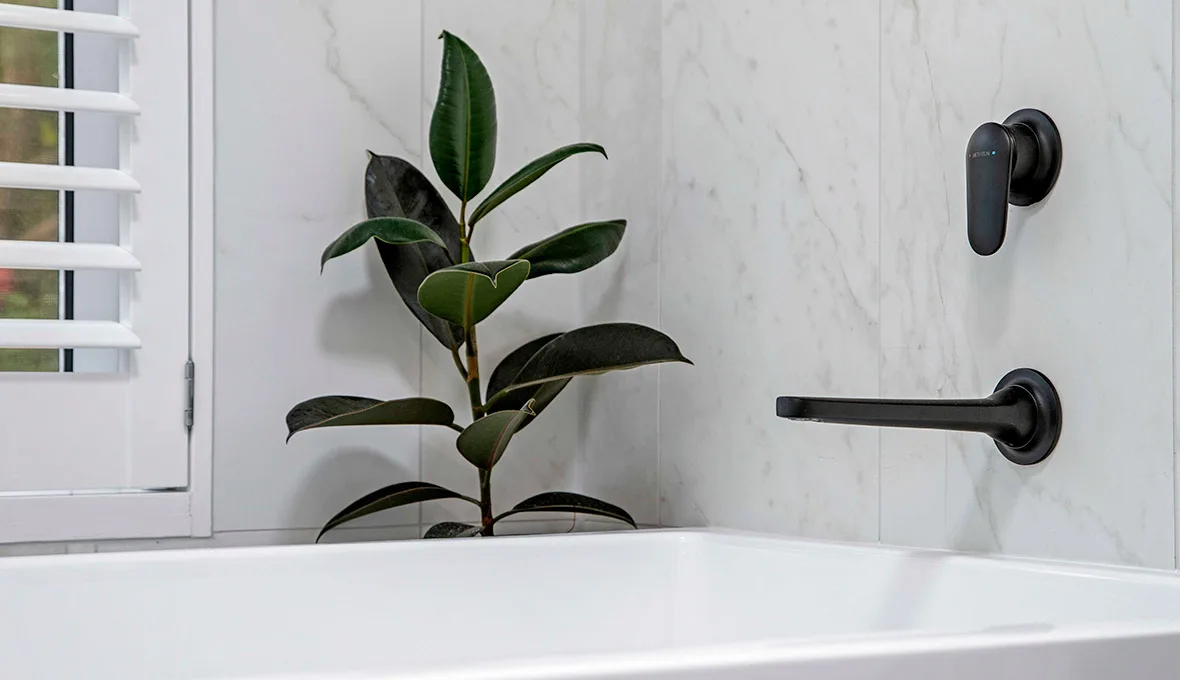 Close-up of a matte black bath spout and mixer on white marble-look tiling, beside a potted rubber plant.