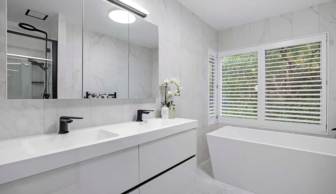 Double vanity with matte black tapware and a round LED mirror reflecting a frameless glass shower, beside a freestanding bath below timber plantation shutters looking onto greenery.
