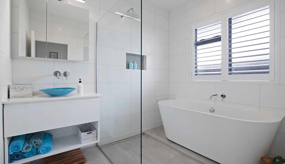 White bathroom with large-format tiles, a frameless glass shower with a rain head, a floating vanity with a turquoise vessel basin, and a freestanding bath below timber plantation shutters.