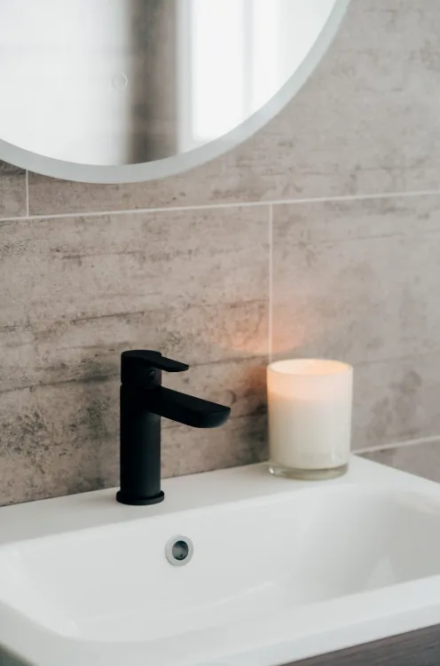 Close-up of a matte black basin mixer on a white rectangular basin, with a lit white candle beside it and the lower edge of a round mirror above.