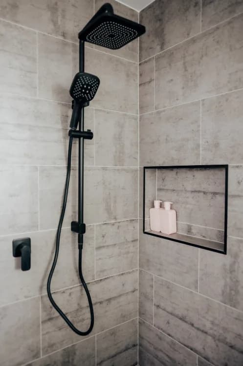 Shower with a matte black square rain head and handheld shower on a riser rail, a black wall mixer, and a black-trimmed recessed niche holding two pale bottles, against warm grey stone-look tiles.