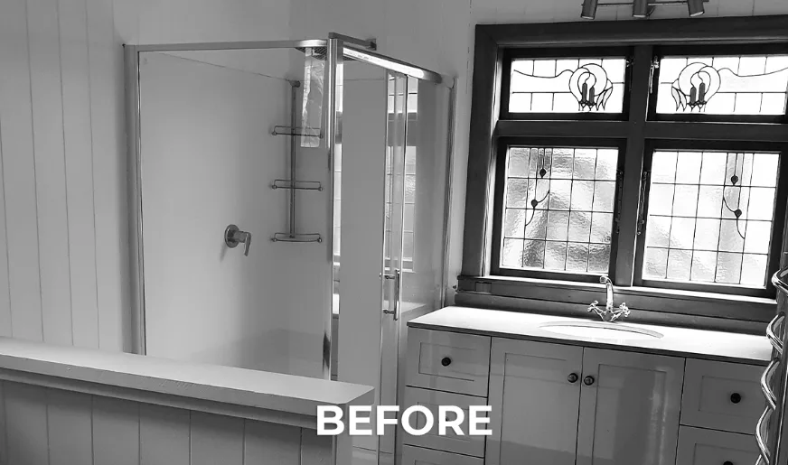 Black-and-white photograph of the bathroom before renovation, showing a chrome-framed shower screen and the same leadlight window with a floral motif above a pedestal vanity.