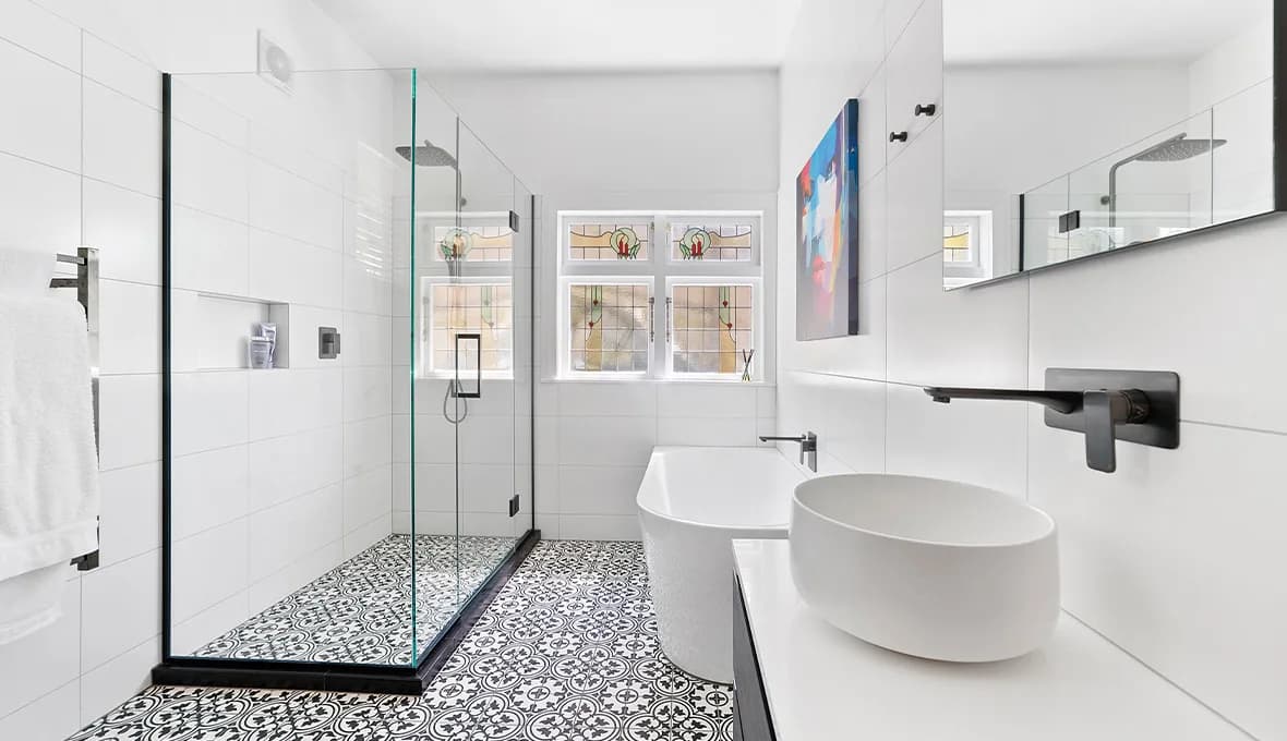 Bathroom with a frameless glass shower and black-and-white patterned floor tiles, an original leadlight window with a floral motif, a colourful portrait artwork on the white tiled wall, and a white freestanding bath with a matte black spout.