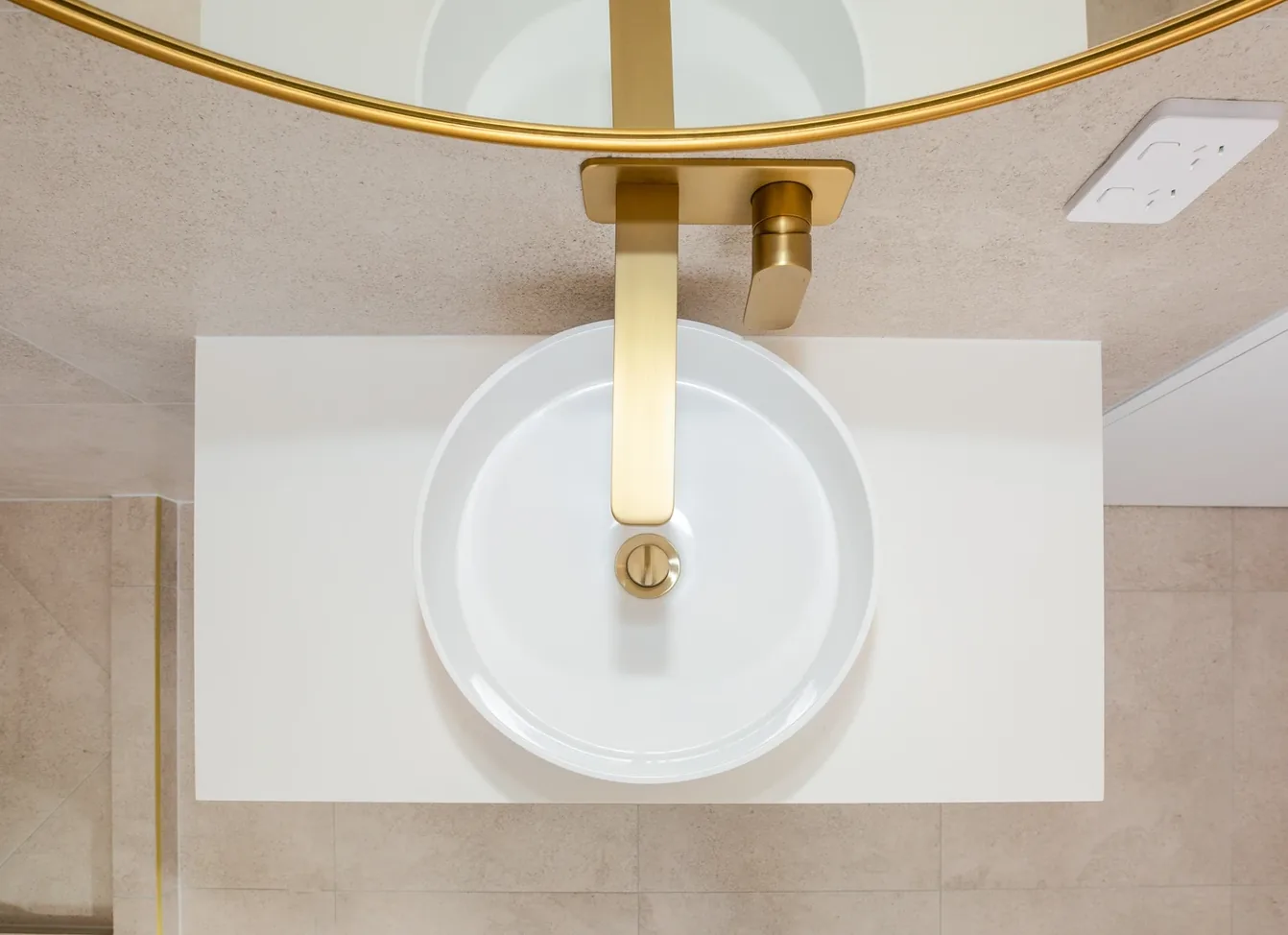 Overhead view of a white round vessel basin on a white benchtop, with a gold wall-mounted spout and mixer and a gold pop-up waste, against sandy beige tiles.