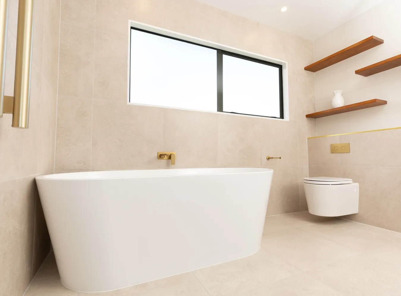 White freestanding oval bath beneath a long horizontal window, with a gold wall-mounted bath spout, beige stone-look floor and wall tiles, and a wall-hung toilet with timber shelves to the right.