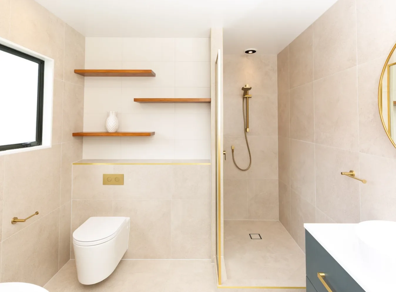 Bathroom finished in sandy beige stone-look tiles, with a walk-in shower behind a gold-trimmed glass screen, a wall-hung toilet with a gold flush plate, and three timber floating shelves set into a white tiled recess.