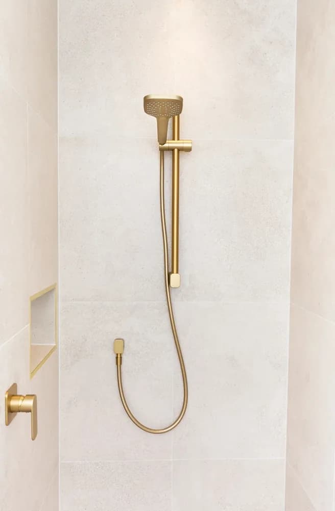 Gold shower rail with a handheld shower head and hose mounted on pale stone-look tiles, with a gold wall mixer and a tiled niche to the left.