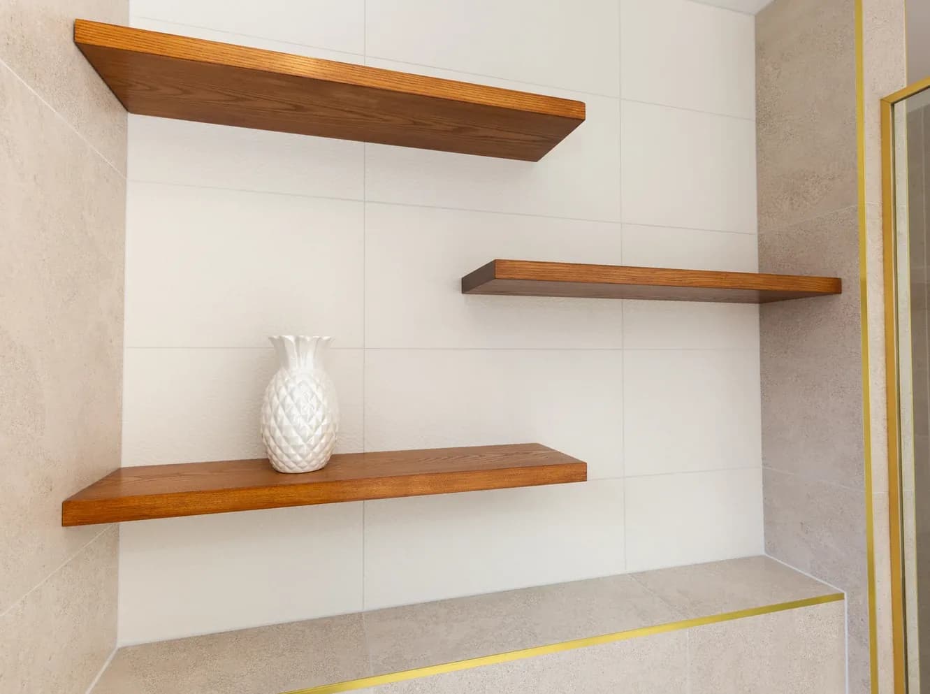 Three staggered solid timber floating shelves mounted in a white-tiled recess, with a white textured pineapple-shaped vase on the lowest shelf.