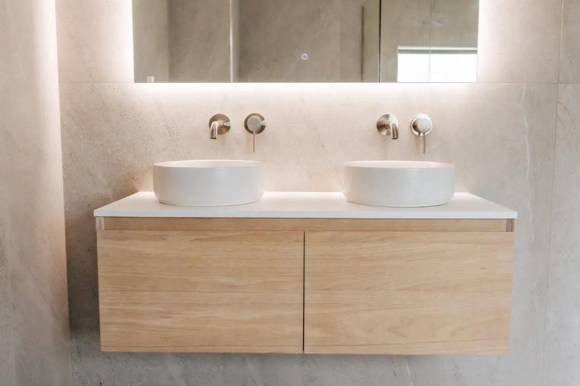 Light oak wall-hung double vanity with a white stone top, two round white vessel basins, brushed nickel wall-mounted mixers, and a wide LED backlit mirror above, against large-format grey stone-look tiles.