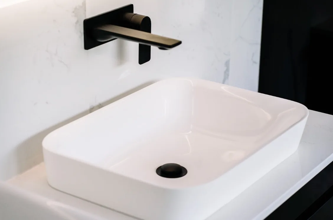 Close-up of a white rectangular vessel basin with a black pop-up waste and a matte black wall-mounted spout and mixer, against white marble-look tiling.