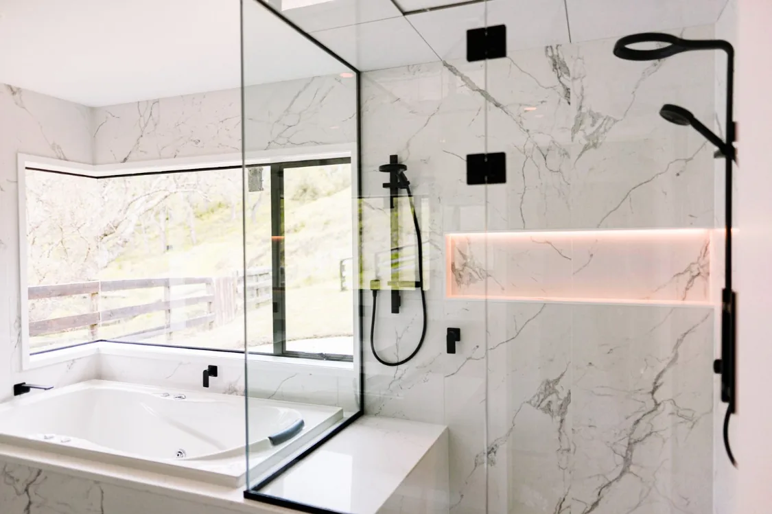 Walk-in shower behind a black-framed glass screen with a black rain head and handheld shower, an LED-lit horizontal niche in the marble-look tiling, and an inset bath beneath a window looking onto trees and a timber fence.