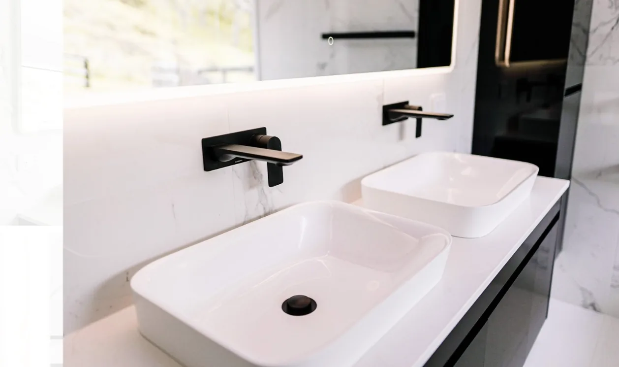 Two white rectangular vessel basins on a white stone top with matte black wall-mounted mixers, an LED backlit mirror above reflecting a window and greenery, and a black gloss cabinet to the right.