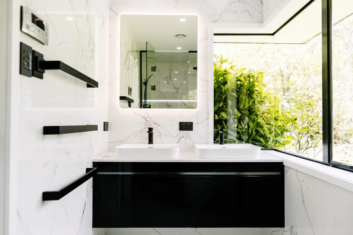 Ensuite with white marble-look tiling, a black gloss wall-hung vanity with a white top and two white vessel basins, matte black tapware, an LED backlit mirror, three black towel rails on the left wall, and a corner window looking onto dense green foliage.