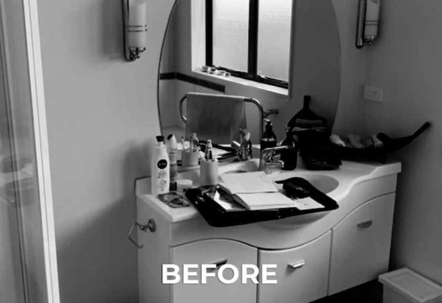 Black-and-white photograph of the original vanity before renovation, cluttered with toiletries and a tray on a curved laminate benchtop, with an oval mirror above.