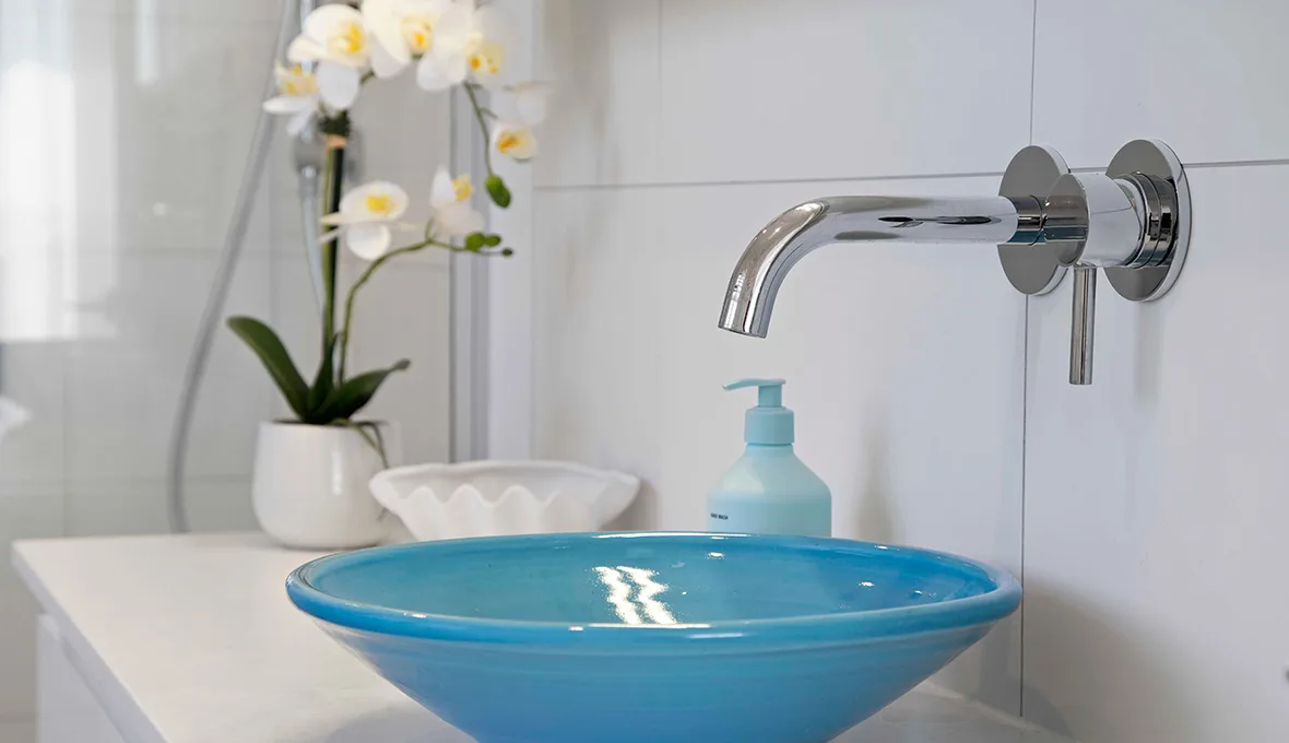 Close-up of a turquoise glass vessel basin with a chrome wall-mounted spout, a white orchid and a shell-shaped dish on the vanity beside it.