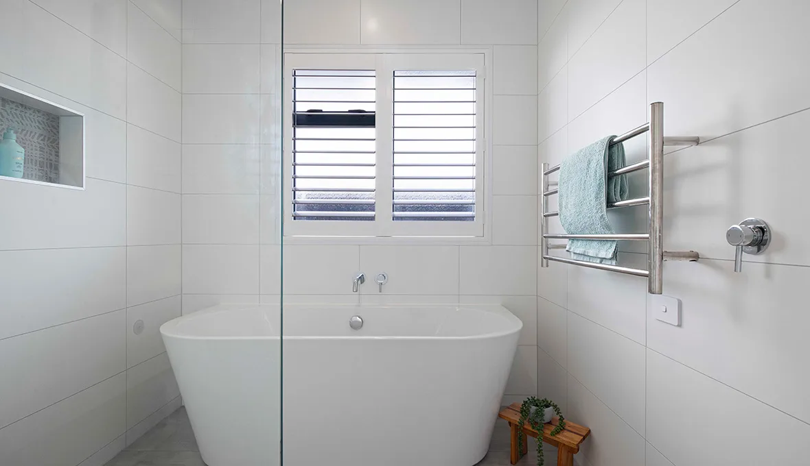 White freestanding bath below timber plantation shutters, with a ladder-style heated towel rail holding a teal towel on the tiled wall beside it.