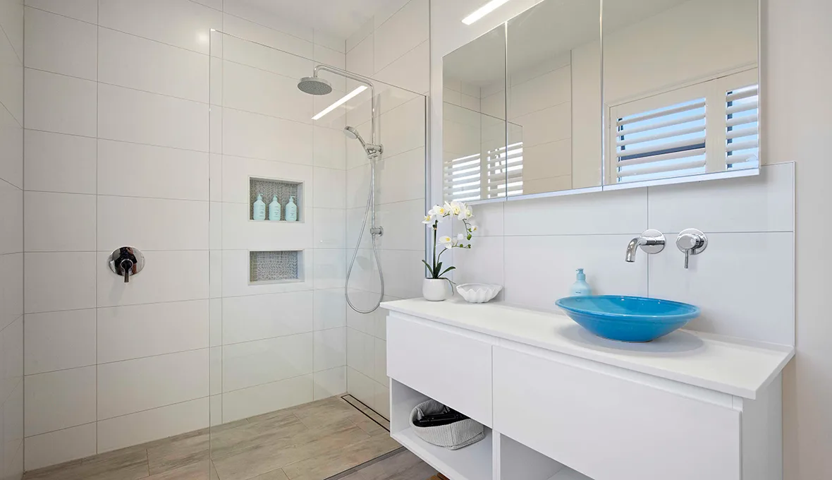 Frameless glass shower with a rain head and tiled niche, beside a wide mirrored cabinet, a floating vanity with a turquoise vessel basin, and timber plantation shutters reflected in the mirror.