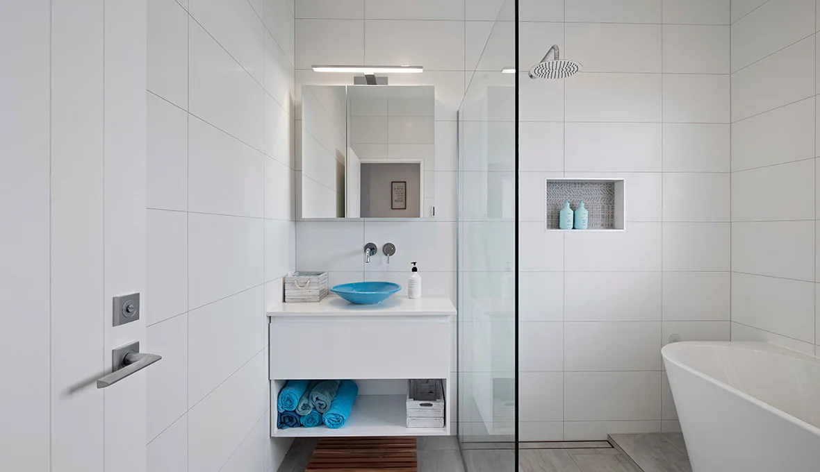 Floating white vanity with a turquoise vessel basin and a chrome wall-mounted tap, folded blue towels on the open shelf below, beside a frameless glass shower with a tiled niche.