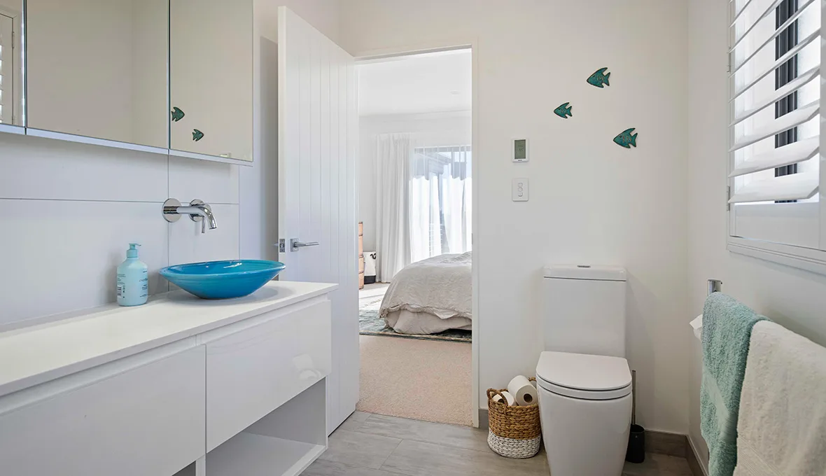 Floating white vanity with a turquoise vessel basin, a close-coupled toilet beside a woven storage basket, a wall-mounted heated towel rail, and an open door leading to an adjoining bedroom.