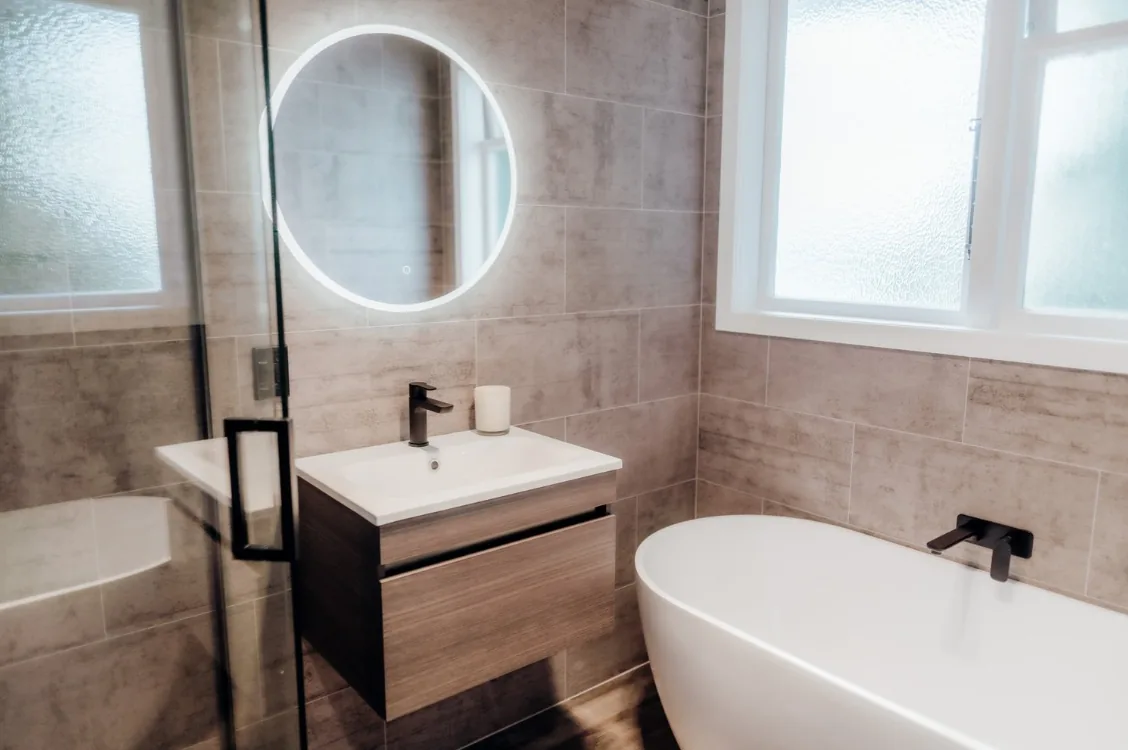 White freestanding bath with a matte black wall-mounted spout beneath an obscured-glass window, next to a timber-look wall-hung vanity with a white basin, black tap and a round LED backlit mirror.