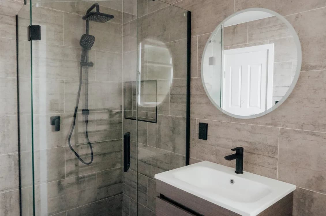 Bathroom in warm grey stone-look tiles with an angled corner shower enclosed by black-framed glass, a black rain head and handheld shower, a timber-look wall-hung vanity with a white basin and black mixer, and a round LED backlit mirror.